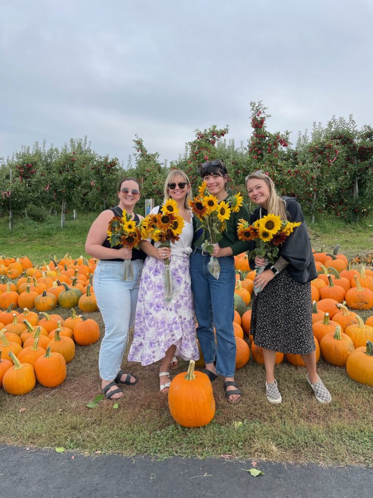 Four friends holding u-pick sunflower bouquets beside rows of pumpkins and apple trees at Chelan Valley Farms in Manson, WA