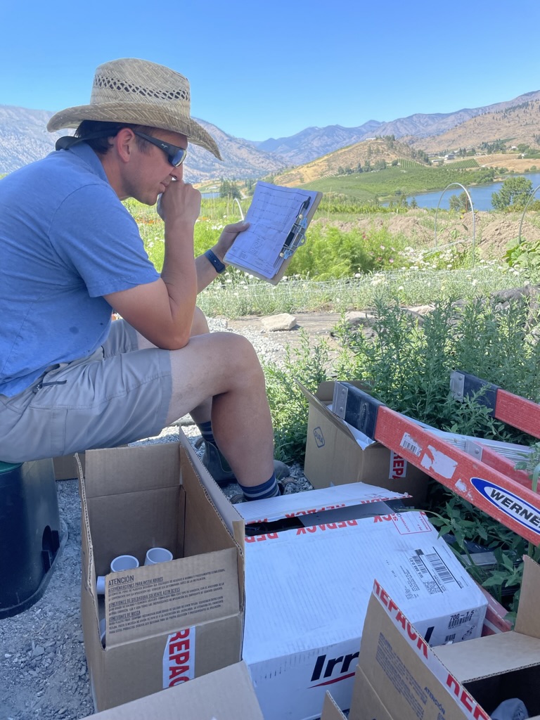 Leadership is lonely - Chad Steiner sitting on an irrigation valve box with a clipboard, planning irrigation at Chelan Valley Farms with Lake Chelan behind him