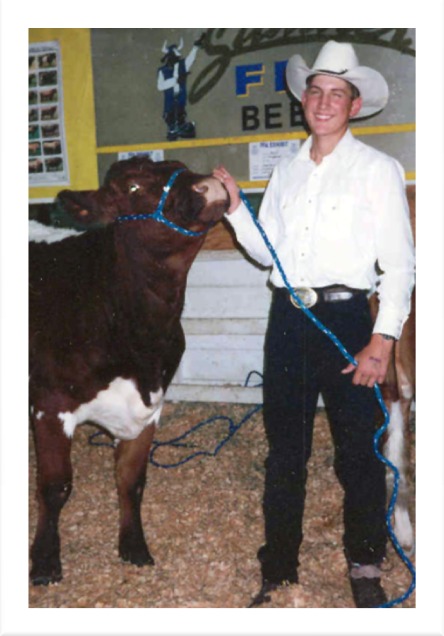 Chad Steiner in a white shirt and cowboy hat showing a red steer in front of the Sumner FFA Beef sign, 1996 – FFA at Chelan Valley Farms